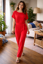 Red short-sleeve jumpsuit with a tie belt, worn by a woman in a living room.