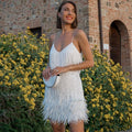 White spaghetti-strap dress with long fringe and feather trim, worn in front of a stone building.