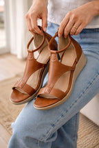 Brown wedge sandals held above jeans to show their straps and footbeds.