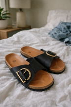 Black slides displayed on a bed with tan footbeds and D-shaped metal details.