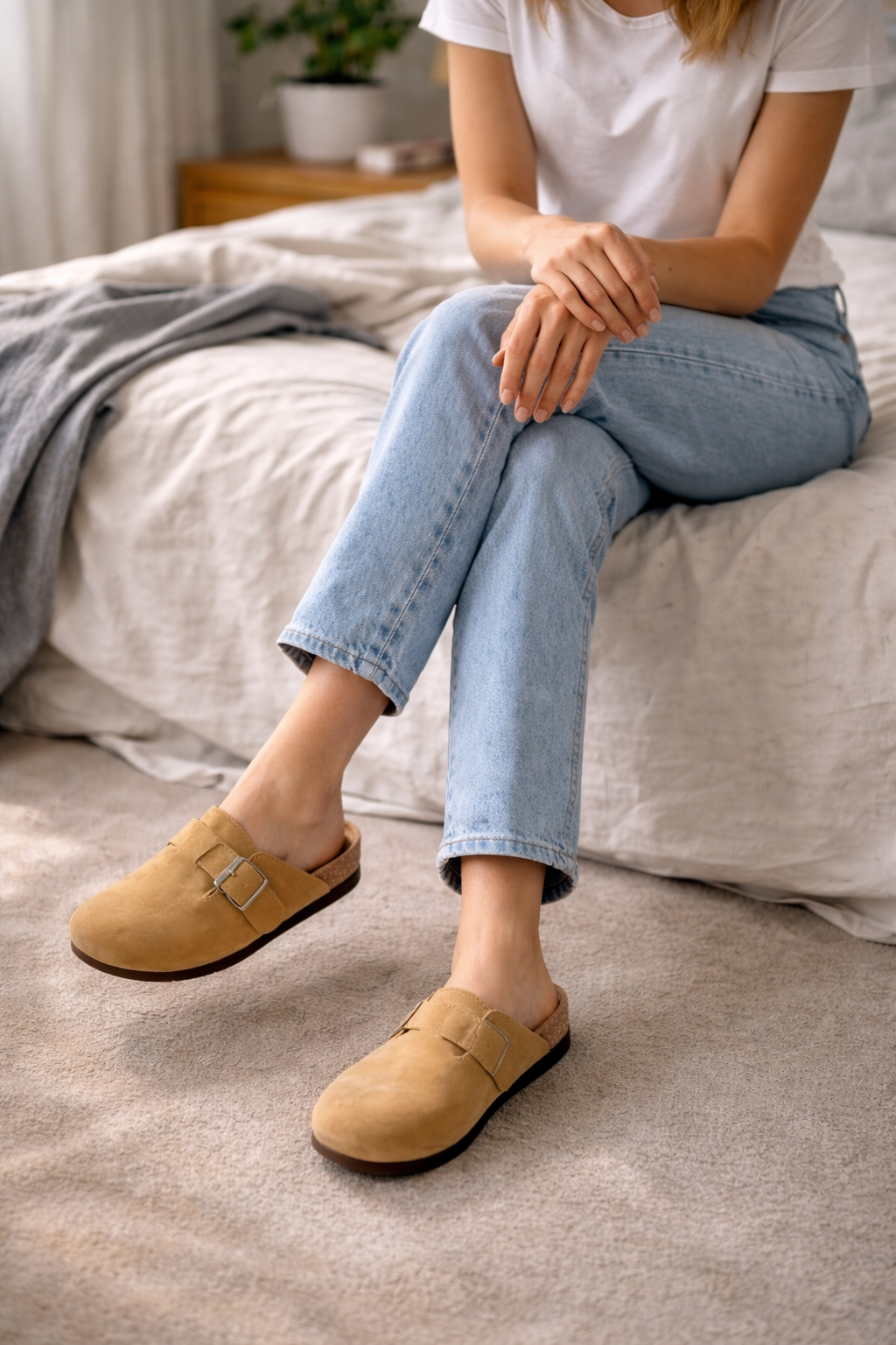 Beige clogs worn by a seated person beside a bed.