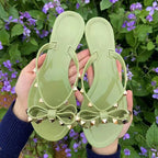 Green sandals with bows and gold-tone studs held against flowers.