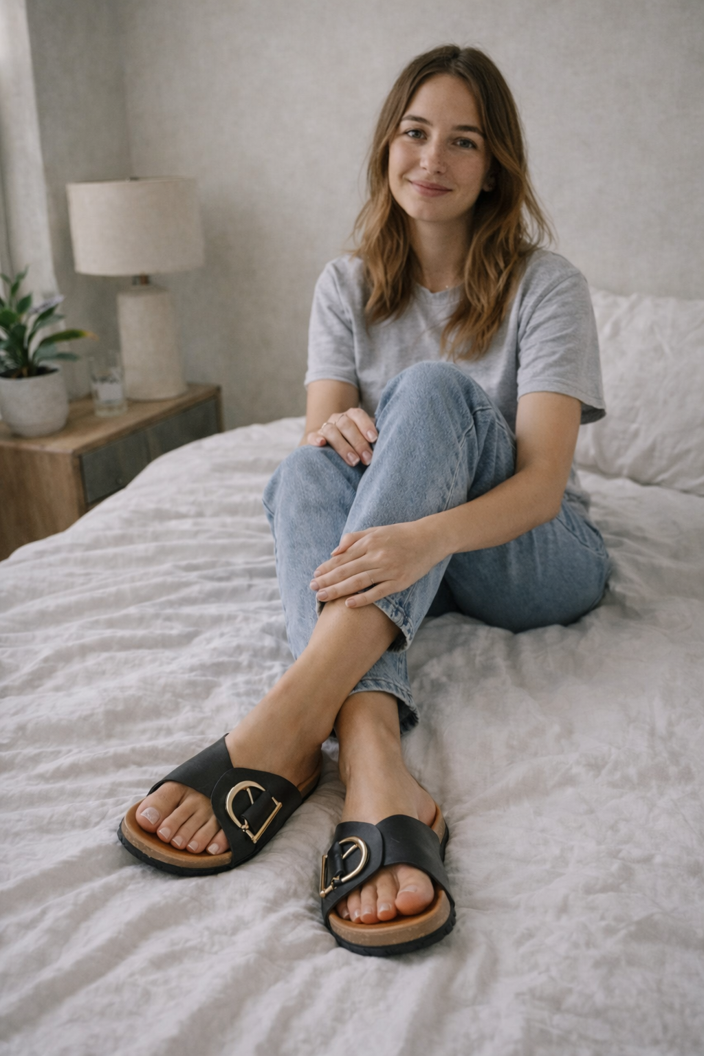 Woman wearing black slides with D-shaped metal details while seated on a bed.