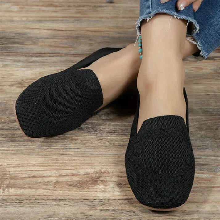 Black slip-on flats with a textured upper, worn with jeans on a wood floor.